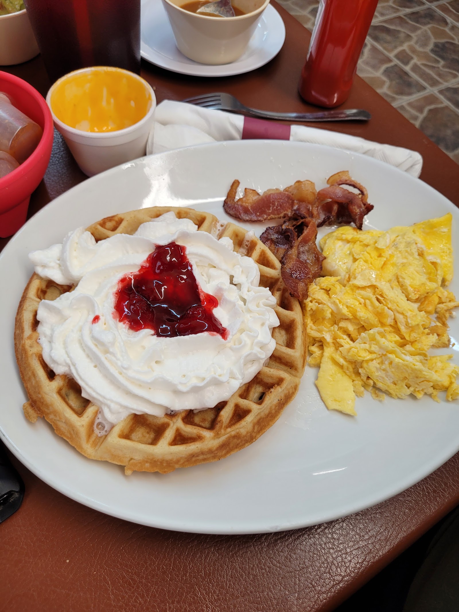 Waffle topped with whipped cream and strawberry sauce, served with eggs and bacon